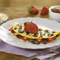 Fotografia em tons de lilás em uma bancada de madeira, com um paninho lilás com bolinhas, um prato branco raso com a crepioca recheada de chocolate e morangos.