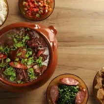 Fotografia vista de cima de uma feijoada completa, com cebolinha por cima, em um recipiente grande clássico de vidro marrom. Ao redor tem acompanhamentos de arroz, torresmo, vingrete e couve, todos em um recipiente de vidro, sobre uma mesa de madeira