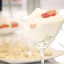 Fotografia de um sorvete de creme com champanhe e raspas de gelo em uma taça. Ao fundo, aperitivos em um recipiente de vidro na cor branco. A granita está sobre uma mesa de madeira na cor branca.