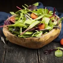 Fotografia em tons escuros de uma salada verde com salsão e tomates cereja dentro de um recipiente fundo de madeira, que está apoiado em um pano azul escuro, juntamente com um garfo de cabo preto, sobre uma mesa de madeira escura.