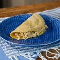 Fotografia em tons de azul com um prato azul ao centro. Em cima do prato existe uma receita de crepioca feita com NESTLÉ Farelo de Aveia e recheada com frango desfiado e creme de ricota