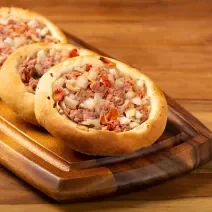 Fotografia de três esfihas de carne, uma apoiada na outra, sobre um apoio de madeira retangular, o qual está apoiado em uma mesa de madeira.