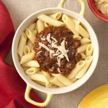 Fotografia em tons de amarelo e vermelho, ao centro uma panela destampada com o macarrão tipo penne e o ragú em cima decorado com queijo ralado. Ao lado está a tampa amarela da panela e um pote vermelho com queijo ralado.