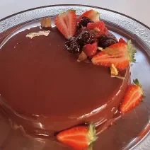 Fotografia em tons de vermelho e marrom com um prato prateado ao centro. Em cima do prato existe um bolo de chocolate coberto com calda de chocolate e frutas vermelhas