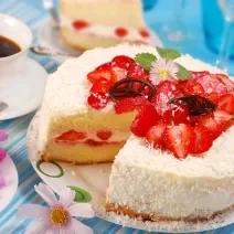 Fotografia de um bolo com recheio e cobertura de coco e morangos, sobre um prato grande de vidro como apoio. Ao redor tem café em uma xícara branca, morangos em um recipiente branco fundo e flores sobre um prato pequeno de vidro branco.