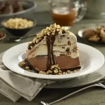 Fotografia de uma fatia de torta Alaska feita de sorvete de café com uma calda de chocolate e amendoins triturados. A torta está em um prato fundo branco.