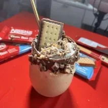 Fotografia de um copo de vidro com a borda com creme de avelã, pedaços de chocotrio, chantilly e uma unidade inteira de chocotrio. Dentro do copo tem milkshake. O copo está sobre uma mesa vermelha, com quatro embalagens de chocotrio.