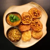 Fotografia vista de cima de seis unidades de pani puri com grão de bico Crocante, e ao lado tem dois recipientes pequenos, um com chutney de cebola e o outro com dahl de lentilha. A receita está em um prato de vidro branco, sobre uma mesa marrom.