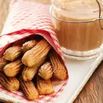 Fotografia em tons de marrom e branco de uma bancada de madeira com um prato retangular, sobre ele um paninho vermelho com churros. Ao lado um recipiente com doce de leite.