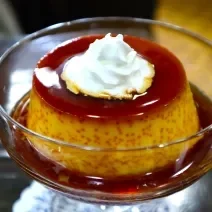 Fotografia de um mini pudim de café dentro de uma taça pequena de vidro com a calda de caramelo. Por cima do pudim tem um pouco de chantilly.