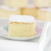 Fotografia em tons claros de um bolo de massa de pão de ló com açúcar de confeiteiro em cima, que está sobre um prato pequeno branco, com desenhos em rosa e azul. Ao fundo, mais pedaços do bolo, e ao lado do prato, colheres de café empilhadas.