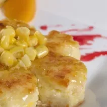 Fotografia de um bolo de milho em pedaços com uma calda e milhos por cima. O bolo está em um prato branco quadrado, com uma calda vermelha no fundo.