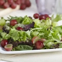 Fotografia de um refratário branco quadrado raso com uma salada de alface, agrião salsão e uva itália com molho de iogurte. O recipiente está apoiado em uma toalha de mesa em tom de bege. Ao fundo tem dois cachos de uva, sendo uvas verdes e uvas roxas.