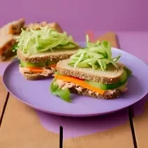Fotografia em tons de roxo com um prato roxo ao centro. Em cima do prato existe dois sanduíches de pão de forma recheados com atum, cenoura e alface.