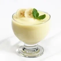 Fotografia de um flan de leite MOÇA e banana dentro de um recipiente de vidro com rodelas de banana e folhas de hortelã. A sobremesa está sobre uma mesa branca.
