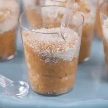 Fotografia em tons de azul com vários potinhos de plástico ao centro. Dentro de cada potinho existe um brigadeiro feito de coco polvilhado com coco ralado.