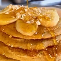 Fotografia cinco panquecas de baunilha empilhadas com mel, banana em rodelas e aveia por cima. As panquecas estão sobre um prato de vidro branco.