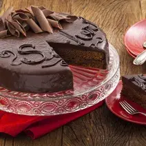 Fotografia em tons de vermelho em uma bancada de madeira com um suporte para bolo de vidro com a torta sacher de chocolate em cima cortada ao meio. Ao lado, pratinhos vermelhos e um deles com uma fatia da torta.