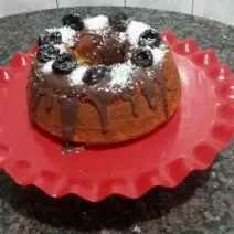 Fotografia de um bolo simples feito na forma de furo central, sobre um apoio grande vermelho decorativo. O bolo está com uma calda de brigadeiro, ameixas e coco ralado por cima.