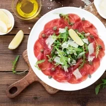 Fotografia vista de cima de fatias de carpaccio com molho de manjericão, nozes, queijo parmesão, folhas verdes e alcaparras.