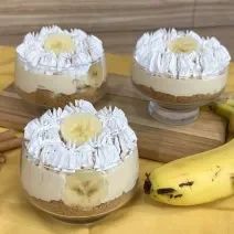 Fotografia em tons de amarelo com um pano amarelo ao centro. Em cima do pano existe uma receita de banoffee feita em pequenos potes de vidro cobertos com chantilly. Ao lado existe duas bananas.