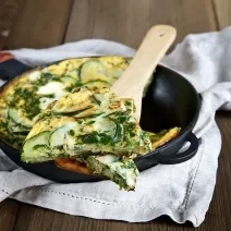 Foto da receita de omelete de abobrinha, servida em uma frigideira preta de ferro, com uma fatia mais próxima, sobre uma colher de pau. A frigideira está sobre um pano bege em uma bancada de madeira.