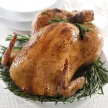 fotografia em tons de branco, verde e bege de uma bancada branca vista de cima. Contém um prato redondo branco com folhas para decorar e por cima um frango assado.