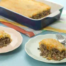 Fotografia em tons de azul em uma bancada de madeira azul, dois pratos redondos rasos com fatias do escondidinho de carne com abobrinha e purê de batata e cenoura em cima de cada um. Ao fundo, um refratário azul escuro com o escondidinho inteiro.