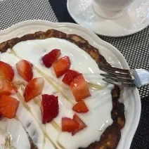 Fotografia de uma panqueca com iogurte, mel e morangos picados por cima sobre um prato raso branco de vidro, ao lado de um garfo e uma xícara branca com café.