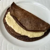 Fotografia de uma panqueca de chocolate com recheio de coco cremoso sobre um prato branco de vidro.