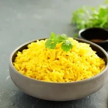 Fotografia em tons de verde com um prato fundo de cerâmica cinza ao centro. Em cima do prato existe um arroz com curry de cor amarelada. Por cima existe uma folha de salsinha.
