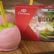 Foto da receita de Maçã verde com cobertura rosa de chocolate. Elas estão sobre uma bancada de madeira com um tecido verde e rosa e uma caixa de KitKat Wicked à direita