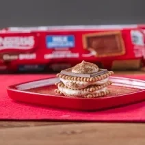 Fotografia de um tiramissu em camadas de creme e choco biscuit sobre um prato quadrado vermelho e raso com cacau em pó polvilhado. O prato está apoiado em um pano vermelho, e ao fundo, tem um pacote de choco biscuit.