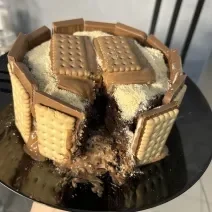 Fotografia de um bolo de chocolate com brigadeiro e farinha láctea polvilhada. Ao redor do bolo tem chocotrio de amendoim grudado. O bolo está sobre um prato de vidro preto raso.