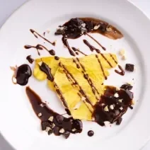 Fotografia vista de cima em tons claros de algumas fatias de abacaxi grelhado com calda de chocolate por cima e ao lado em um prato branco, que está apoiado sobre uma mesa branca.
