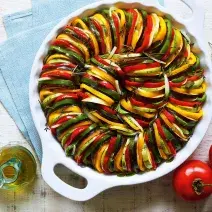 Fotografia em tons de verde, amarelo e vermelho de uma bancada branca com três tomates, uma jarrinha de azeite, ramos de alecrim e um recipiente branco redondo com alças com camadas intercaladas de abobrinha, berinjela e pimentões.