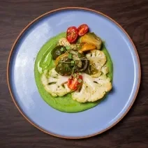 Fotografia em tons de azul com um prato azul ao centro. Em cima do prato existe uma receita de couve-flor assada acompanhada por salada de jiló com tomates cereja e um molho de cor verde.