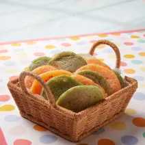 Fotografia em tons de verde e laranja em uma bancada de madeira, uma toalha branca com bolinhas coloridas, uma cesta de vime marrom clara com pães de queijo achatados nas cores verde e amarelo.