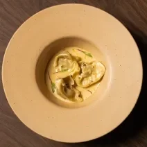 Foto da receita de Ravioli de Escargot feito no Masterchef 11. Observa-se um prato fundo visto de cima com três raviolis servidos com molho e decorado com brotos.