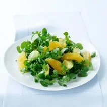 Fotografia em tom claro, um prato branco com uma salada de agrião e pedaços de tangerina, sobre uma toalha de mesa azul em tom claro.
