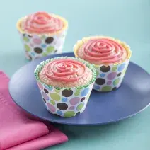 Fotografia em tons de azul e rosa em uma mesa de madeira azul, um pano rosa, um prato azul redondo com dois cupcakes de morango com aveia em forminhas coloridas.