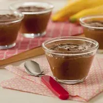 Fotografia em tons de vermelho em uma bancada de madeira com paninhos desenhados em rosa e vermelho e uma tábua de madeira com dois potinhos com mousse de cacau e abacate em cima dela. Ao centro, um potinho com o mousse e uma colher com cabo vermelho.