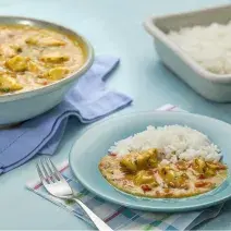 Fotografia em tons de azul e amarelo, ao centro prato azul com frango ensopado e arroz, sobre guardanapo azul e branco com um garfo, à direita travessa com arroz, à esquerda travessa com frango ensopado e guardanapo azul, tudo sobre bancada na cor azul.