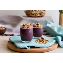 Fotografia de dois copos de vidro, os dois com shake de açaí e granola por cima. Os copos estão sobre um pano azul de tom claro, com mais granola, no canto. O pano está sobre uma tábua redonda de madeira.