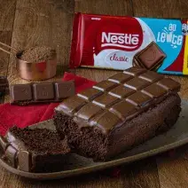 Foto da receita de Chocolate Bar Cake, sobre uma bancada de madeira, em um prato em tom marrom. Na bancada há uma barra de chocolate ao leite, um copo de medida de cobre com cacau em pó e alguns pedaços de chocolate