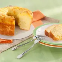 Fotografia em tons de laranja e verde em uma mesa de madeira verde com um pano laranja de bolinhas branca. Ao centro, um prato raso branco com o bolo de macaxeira cortado. Ao lado, um prato branco com borda verde e duas fatias do bolo.