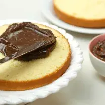 Foto de um bolo aberto ao meio em cima de um prato branco sendo preenchido com creme de chocolate por cima