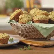 Fotografia em tons de verde em uma bancada de madeira, uma tábua de madeira, uma cesta de vime com vários muffins de aveia e embaixo um paninho verde. Ao lado, um pratinho de sobremesa branco com um muffin em cima dele.