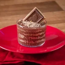 Fotografia de um copo de vidro pequeno com camadas de chocolate alpino derretido e chocobiscuit triturado. Por cima tem choco biscuit triturado e um biscoito inteiro apoiado para decorar. O copo está sobre um prato e uma toalha de mesa vermelha.