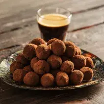 fotografia em tons de marrom e preto de uma bancada marrom vista de frente, contém um prato redondo com trufas com chocolate, e ao fundo um copo com café.